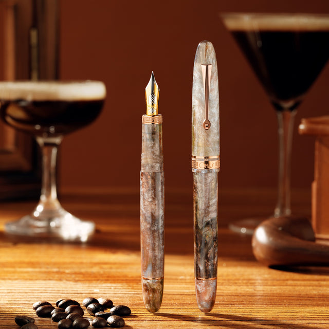 Two fountain pens on a wooden surface with coffee beans and a glass of dark liquid in the background.