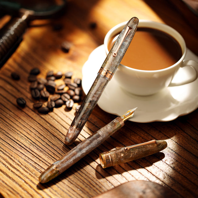 Two vintage-style pens on a wooden surface with a cup of coffee and coffee beans.