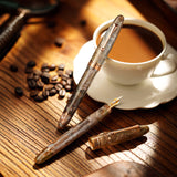 Two vintage-style pens on a wooden surface with a cup of coffee and coffee beans.